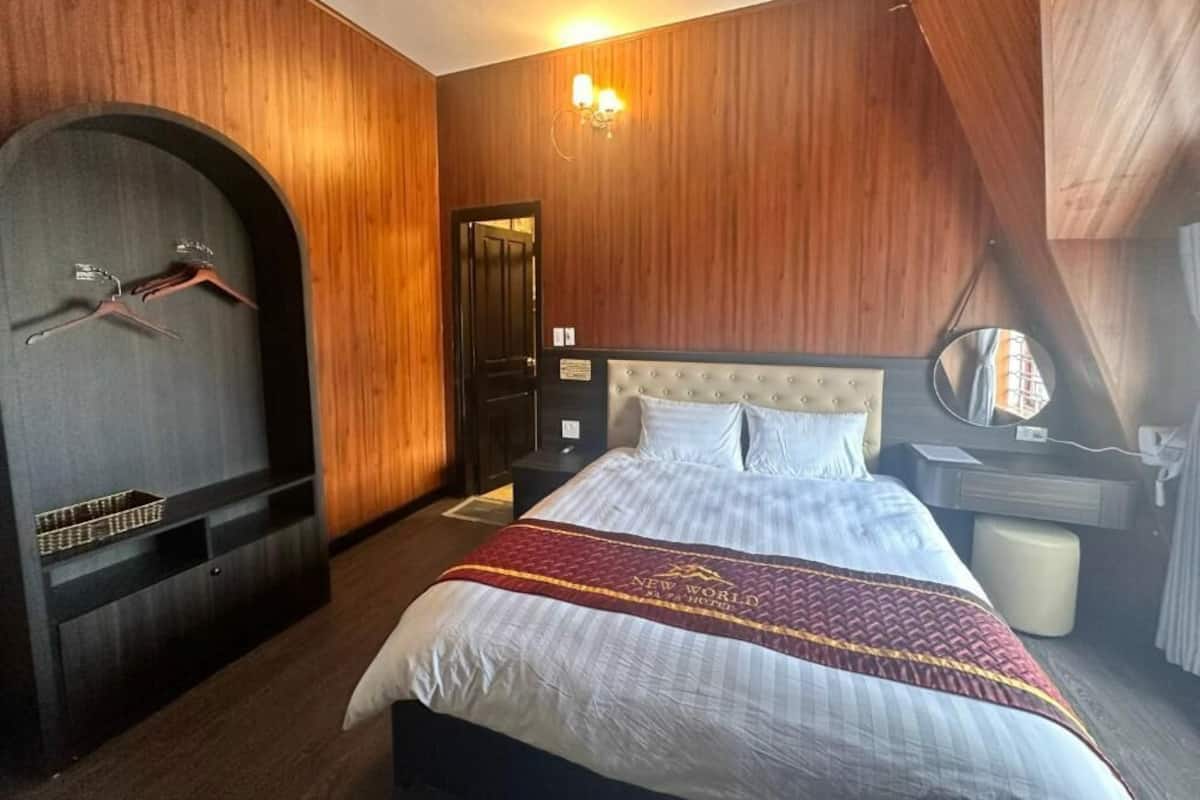 A hotel room with a large bed, wooden paneling, a wardrobe with hanging clothes, a bedside table, and a mirror.