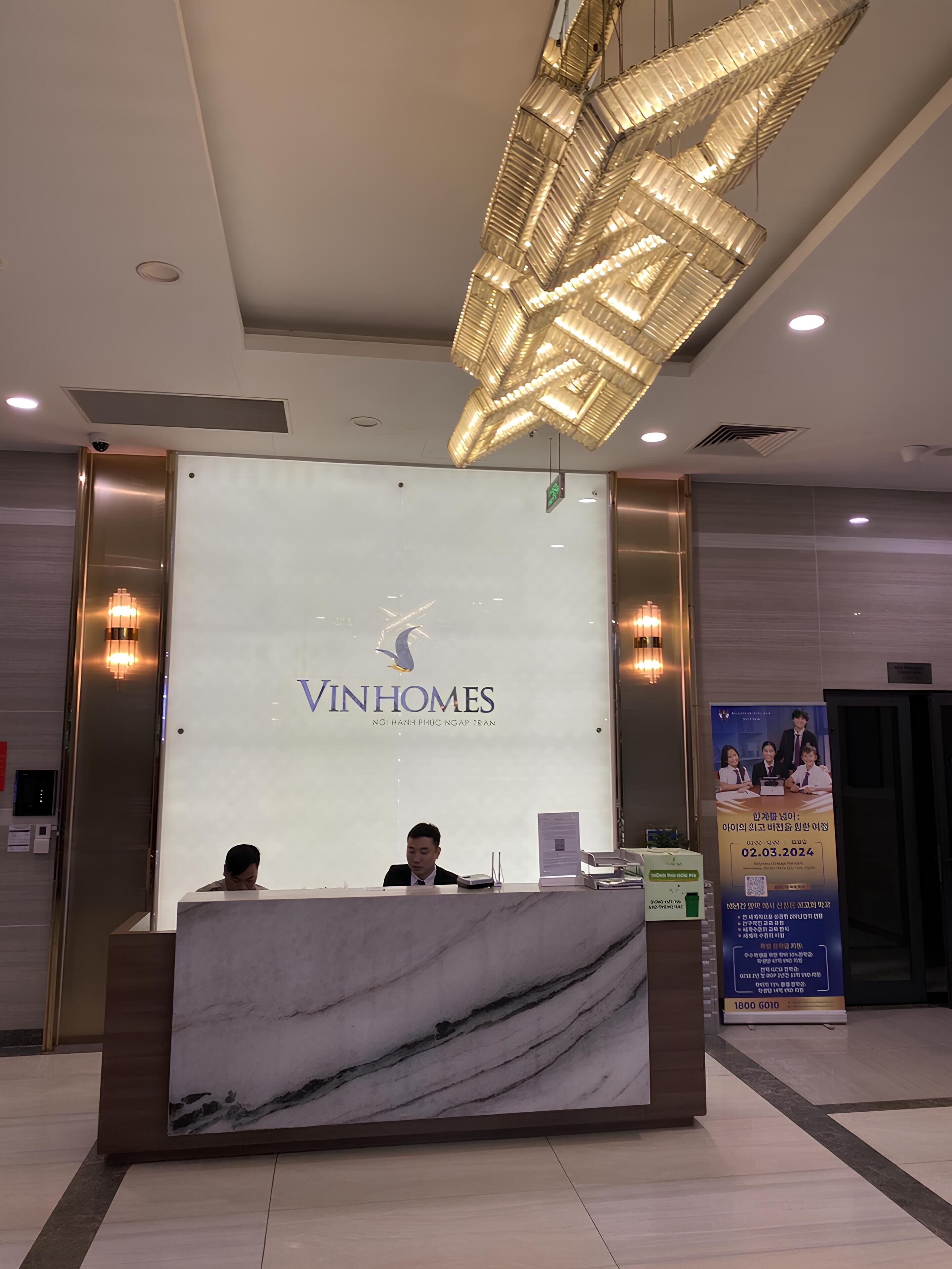 A reception desk with a marble top, a large illuminated sign reading "VINHOMES," and a modern chandelier above.