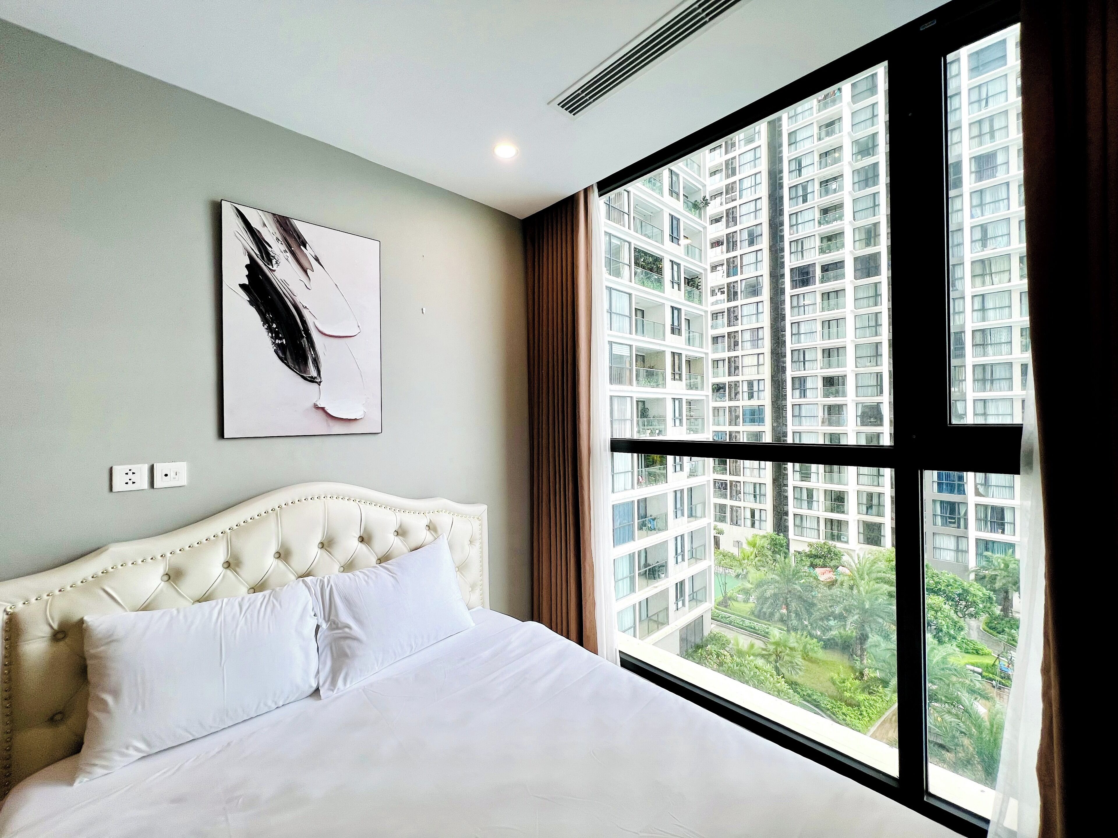 Habitación de hotel con un ventanal grande, cama con ropa blanca, cabecera capitonada y un cuadro en la pared.