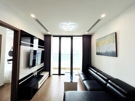 Un salón moderno con televisor de pantalla plana, un sofá de cuero negro, un cuadro en la pared y vistas de la ciudad a través de amplios ventanales.