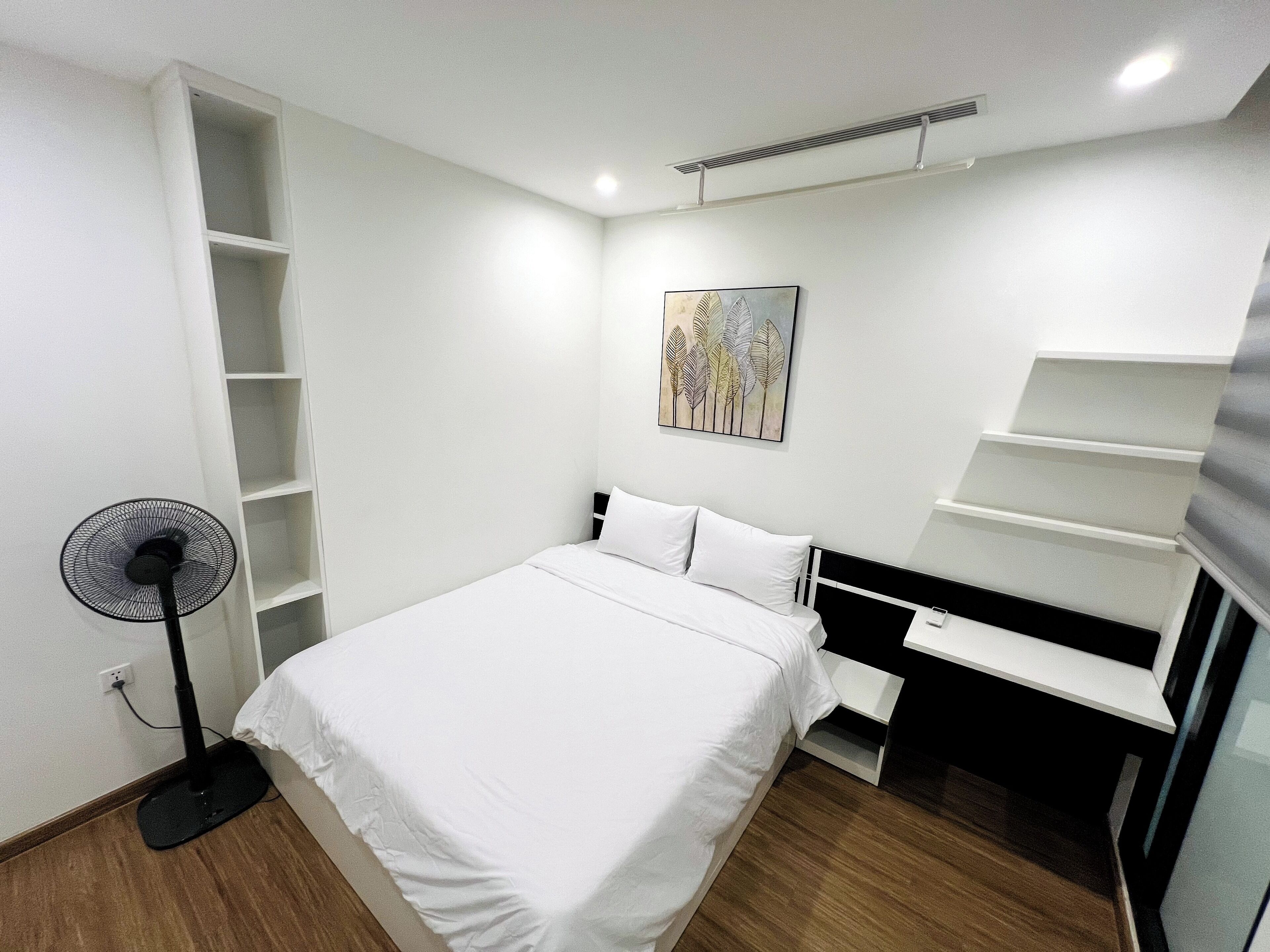 Een slaapkamer met een bed, een ventilator, een bureau en een wandplank.