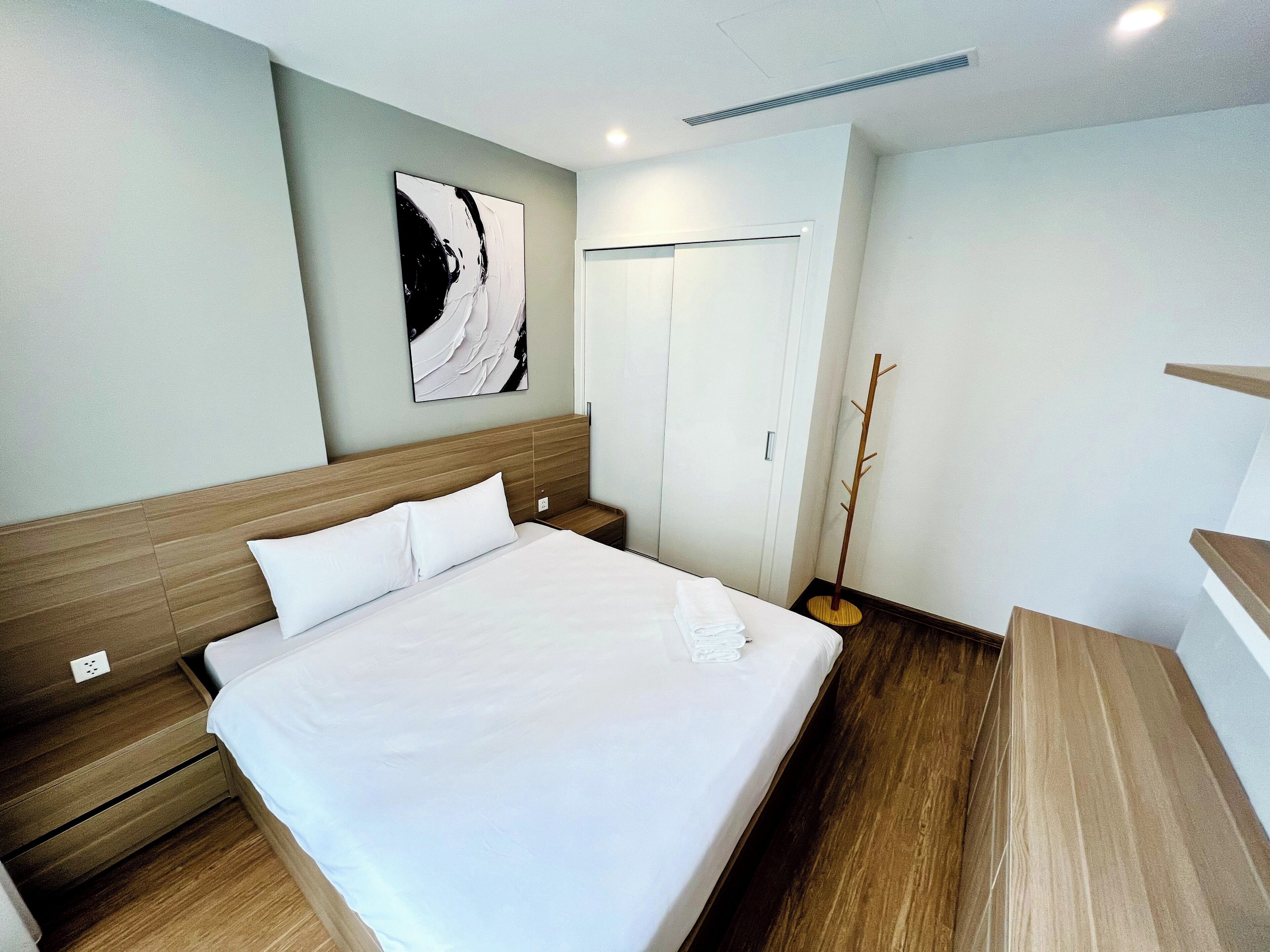 Quarto de hotel com uma cama grande, cabeceira de madeira, um cabideiro e uma pintura abstrata pendurada na parede.