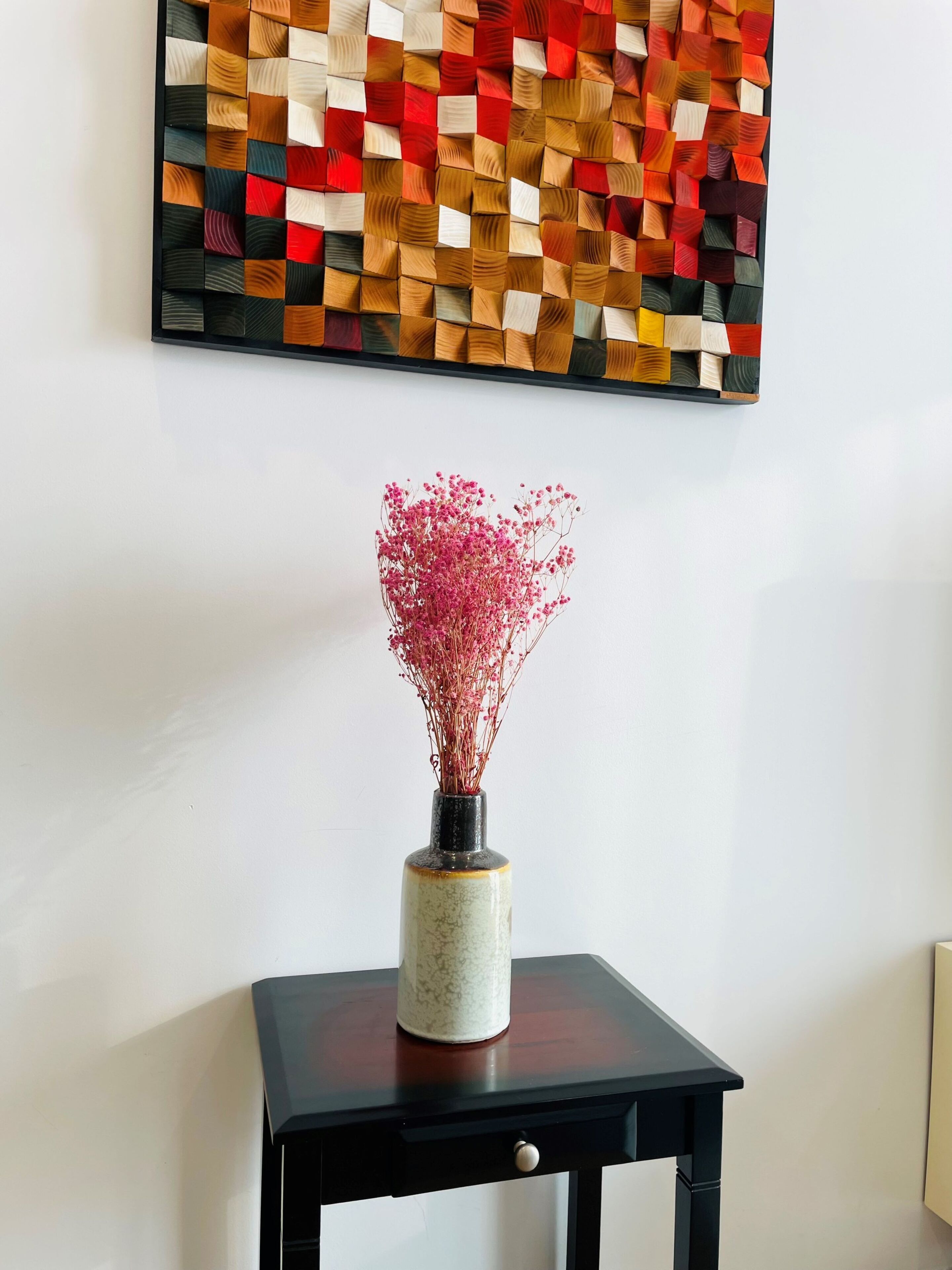 A vase with pink flowers on a black table against a white wall with a colorful abstract art piece above.