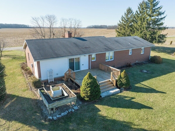 A two-story house with a front deck, a small garden, and a large field in the background.