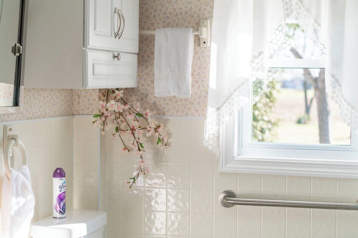 Ein Badezimmer mit Toilette, Waschbecken, Spiegel, Handtuchhalter, einem Fenster mit Spitzenvorhängen und einer Blumendekoration.