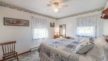 A bedroom with a bed, a chair, a dresser, a ceiling fan, and two windows with sheer curtains.
