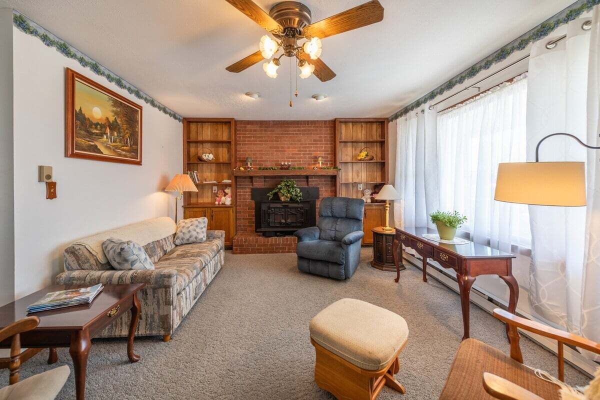 Un salon avec une cheminée, un ventilateur de plafond, un canapé, un fauteuil, une table et une fenêtre avec des rideaux.