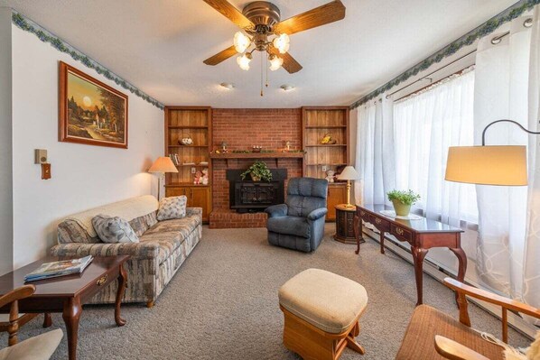 A living room with a fireplace, a ceiling fan, a sofa, a chair, a table, and a window with curtains.