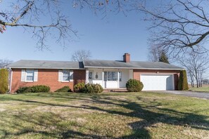 A single-story brick house with a white garage door, a chimney, and a small front yard with trimmed bushes.