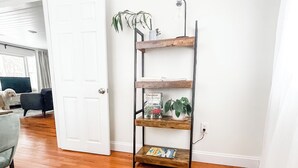 Ein Wohnzimmer mit Holzboden, einem gewebten Teppich, einem Metallregal mit Pflanzen und einer Tür zu einem anderen Raum.