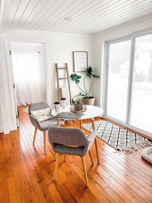 Un salon moderne avec un plancher en bois, des murs blancs et une grande fenêtre. On y trouve une table blanche sur laquelle se trouve un ordinateur portable, une plante en pot et, accrochée au mur, une citation encadrée.