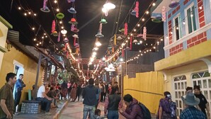 A corridor with colorful hanging decorations and string lights, leading to a building with a yellow facade and a red brick wall. - Casa chikuwa  (San Juan La Laguna)