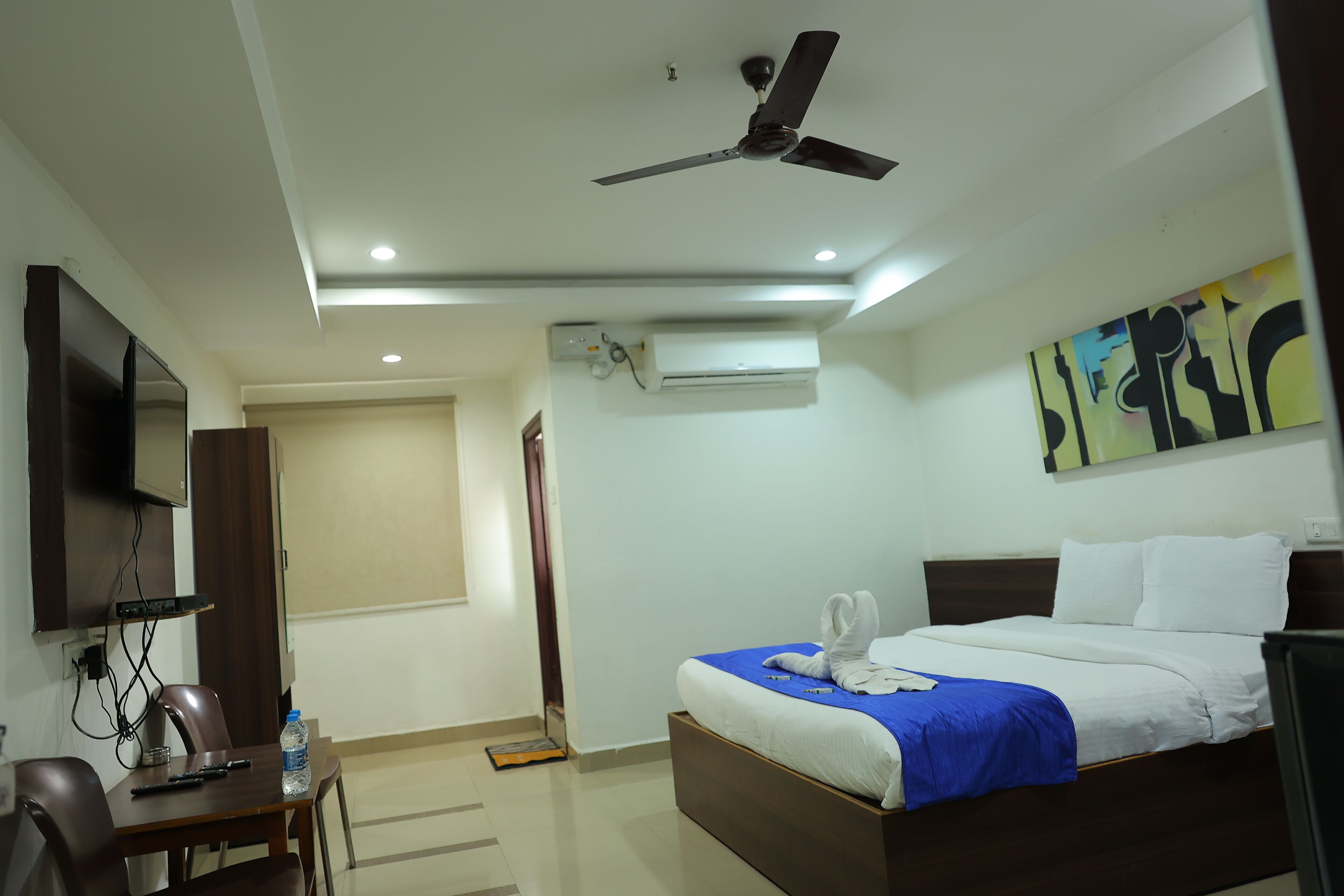 A hotel room with a bed, a desk, a chair, a ceiling fan, an air conditioning unit, and a painting on the wall.