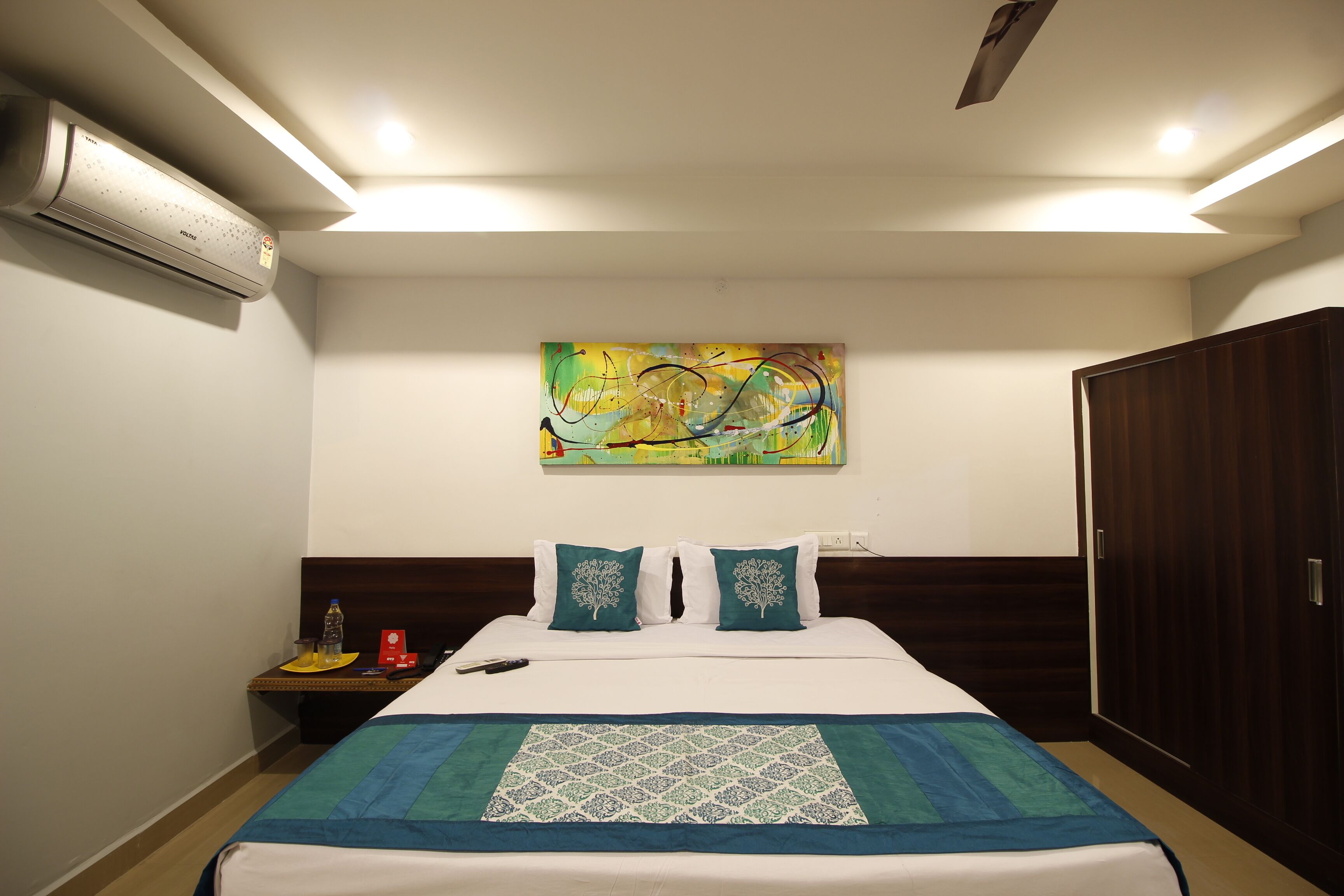 A hotel room with a bed, a nightstand, an air conditioning unit, a painting on the wall, and a wardrobe.