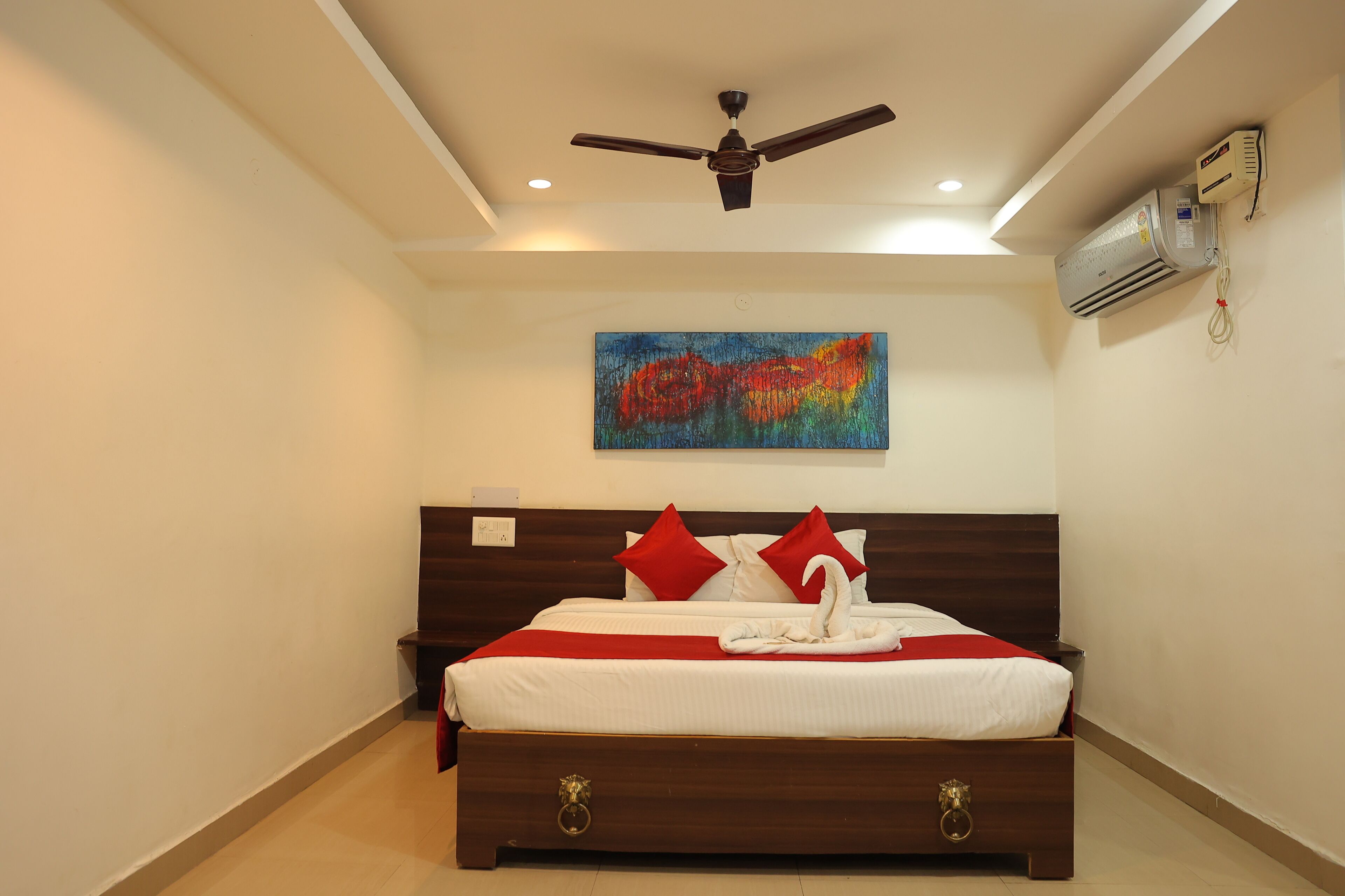 A bedroom with a bed, a ceiling fan, an air conditioning unit, and a colorful abstract painting on the wall.