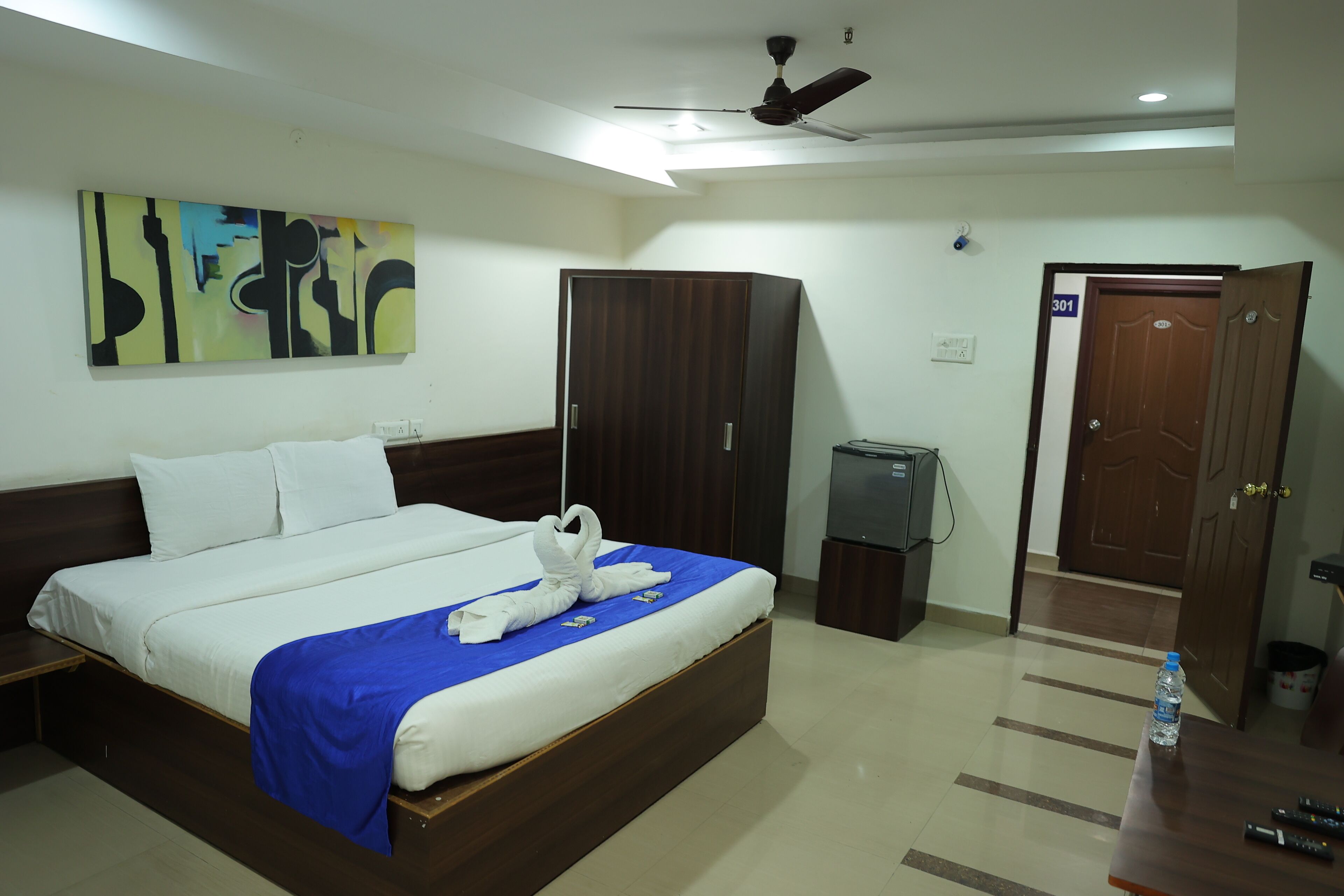 A hotel room with a bed, a wardrobe, a desk with a water bottle, and a door with a number.