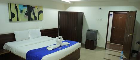 A hotel room with a bed, a wardrobe, a desk with a water bottle, and a door with a number.