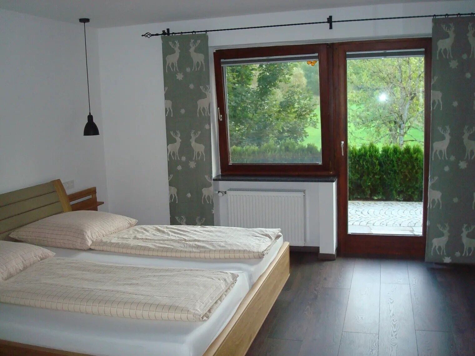 Ein Schlafzimmer mit zwei Betten, einem Fenster mit Blick auf Grünflächen, einem Heizkörper und einer Pendelleuchte.
