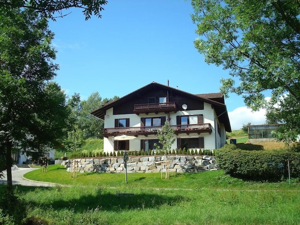 A two-story chalet with a stone foundation, a balcony with wooden railings, and a clear sky. - Apartment 'Ferienwohnungen Memminger' with Private Terrace and Wi-Fi (Aicha vorm Wald)