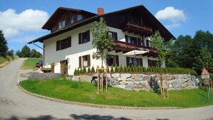 A two-story house with a stone foundation, a balcony with a wooden railing, and a driveway leading to a parked car. - Apartment 'Ferienwohnungen Memminger' with Private Terrace and Wi-Fi (Aicha vorm Wald)