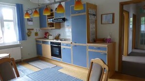 Una cocina funcional con armarios azules, estufa y lavabo. Hay sillas de madera y una mesa con mantel a cuadros.