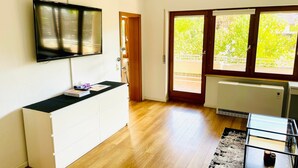 Un salon moderne avec un téléviseur à écran plat fixé au mur, un meuble blanc, une table basse en verre et une vue dégagée sur l’extérieur grâce à une grande porte-fenêtre coulissante.