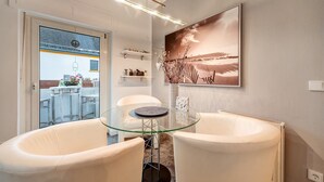 Un intérieur moderne avec une table en verre, des chaises blanches, un grand tableau représentant un paysage et une vue à travers une porte vitrée.