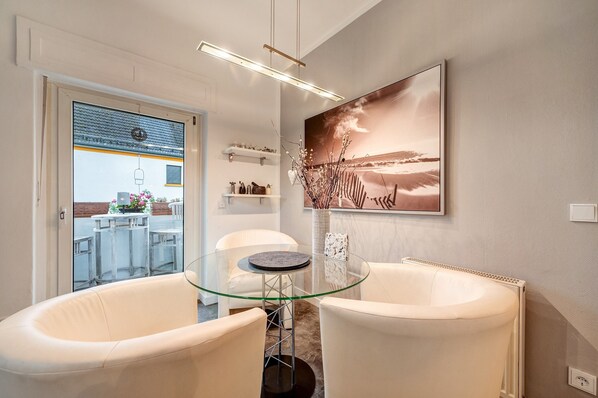 Un intérieur moderne avec une table en verre, des chaises blanches, un grand tableau représentant un paysage et une vue à travers une porte vitrée.