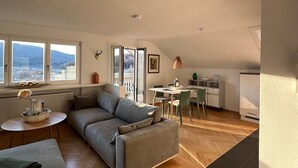 A modern loft apartment with a herringbone wood floor, a gray sofa, a dining area, and a kitchen with a view.