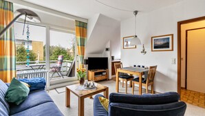 A modern living room with a sofa, coffee table, TV, dining table, chairs, and a balcony with a view. - Apartment 'Utspann' with Balcony and Wi-Fi (Sankt Peter-Ording)