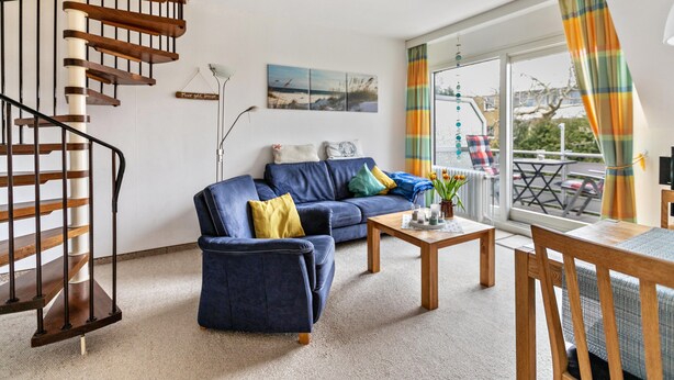 A living room with a blue sofa, a wooden coffee table, a spiral staircase, and a large window with curtains. - Apartment 'Utspann' with Balcony and Wi-Fi (Sankt Peter-Ording)