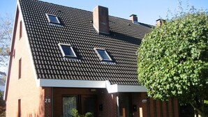 A two-story brick house with a sloped roof, three windows, and a small front yard with a tree and shrubs. - Apartment 'Utspann' with Balcony and Wi-Fi (Sankt Peter-Ording)