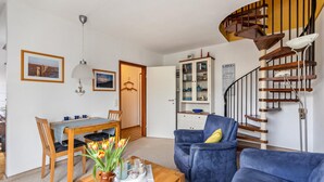 A living room with a wooden dining table, blue armchairs, a blue sofa, a spiral staircase, and a kitchen cabinet. - Apartment 'Utspann' with Balcony and Wi-Fi (Sankt Peter-Ording)