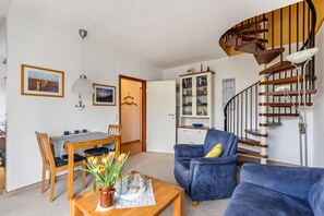 A living room with a wooden dining table, blue armchairs, a blue sofa, a spiral staircase, and a kitchen cabinet.