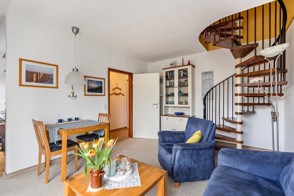A living room with a wooden dining table, blue armchairs, a blue sofa, a spiral staircase, and a kitchen cabinet.