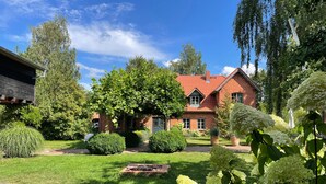 A red brick house with a well-maintained lawn, a wooden deck, and a variety of greenery including bushes and trees.