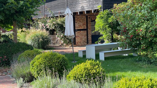 A garden with a bench, a white umbrella, and a brick building.