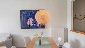 Een moderne eetruimte met een tafel, stoelen, een vaas met bloemen, een hanglamp en een schilderij aan de muur.