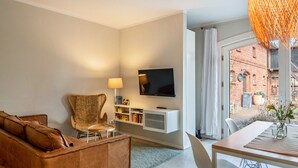 A modern living room with a sofa, a chair, a TV mounted on the wall, a dining table with chairs, and a view of a brick building through large windows. - Holiday Apartment DREI at Hof Himmelgrün with Large Garden (Saal)