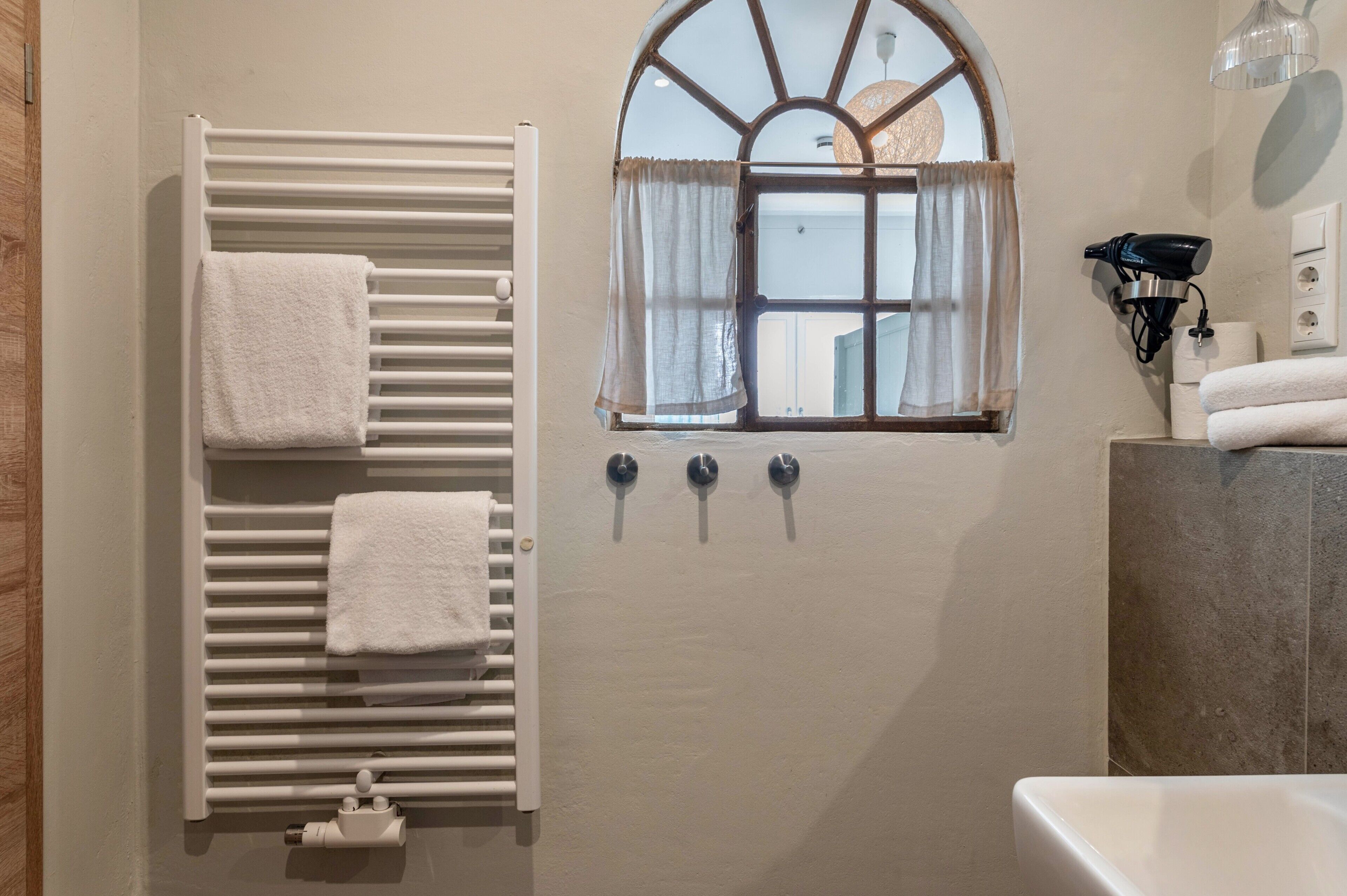 A bathroom with a towel rack, a window with curtains, a wall-mounted hairdryer, and a bathtub.