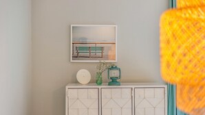 Un mur orné d’un tableau représentant un banc au bord de la mer, un meuble blanc avec des tiroirs et un petit vase vert contenant des fleurs.