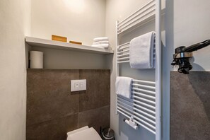 Een badkamer met een handdoekrek, een plankje met handdoeken, een toilet en een prullenbak.
