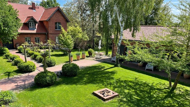 Ett hus byggt av tegel med ett rött tak, omgivet av en välskött trädgård med olika växter och en eldstad.