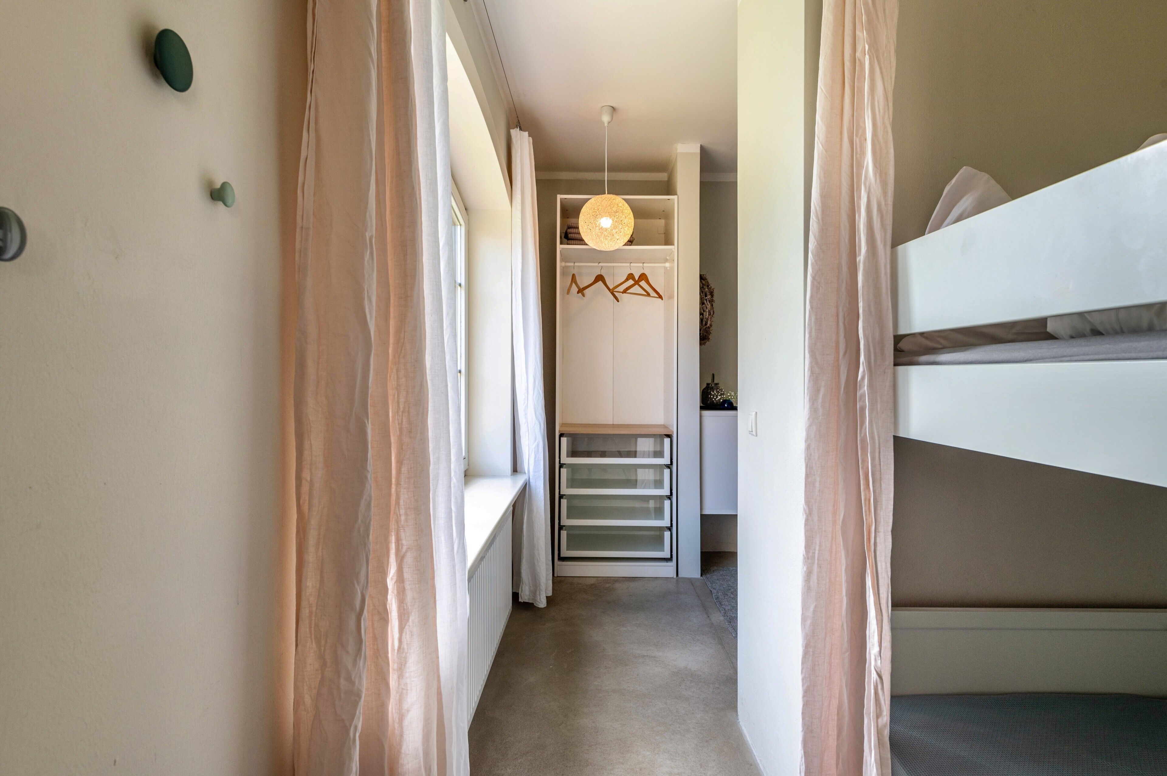 Un couloir étroit mène à une chambre où se trouve un lit superposé, un luminaire suspendu et un placard équipé d’une tringle à vêtements en bois.