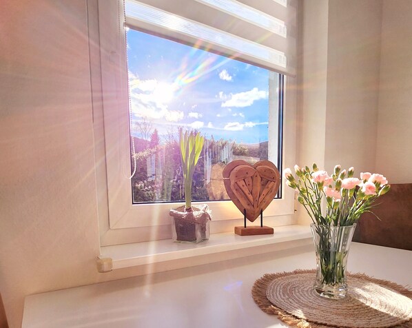 A window with a view of a landscape, a heart-shaped wooden decoration, and a vase with flowers on a table.