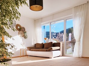 A modern living room with a white sofa, a large window with curtains, and a hanging light fixture.