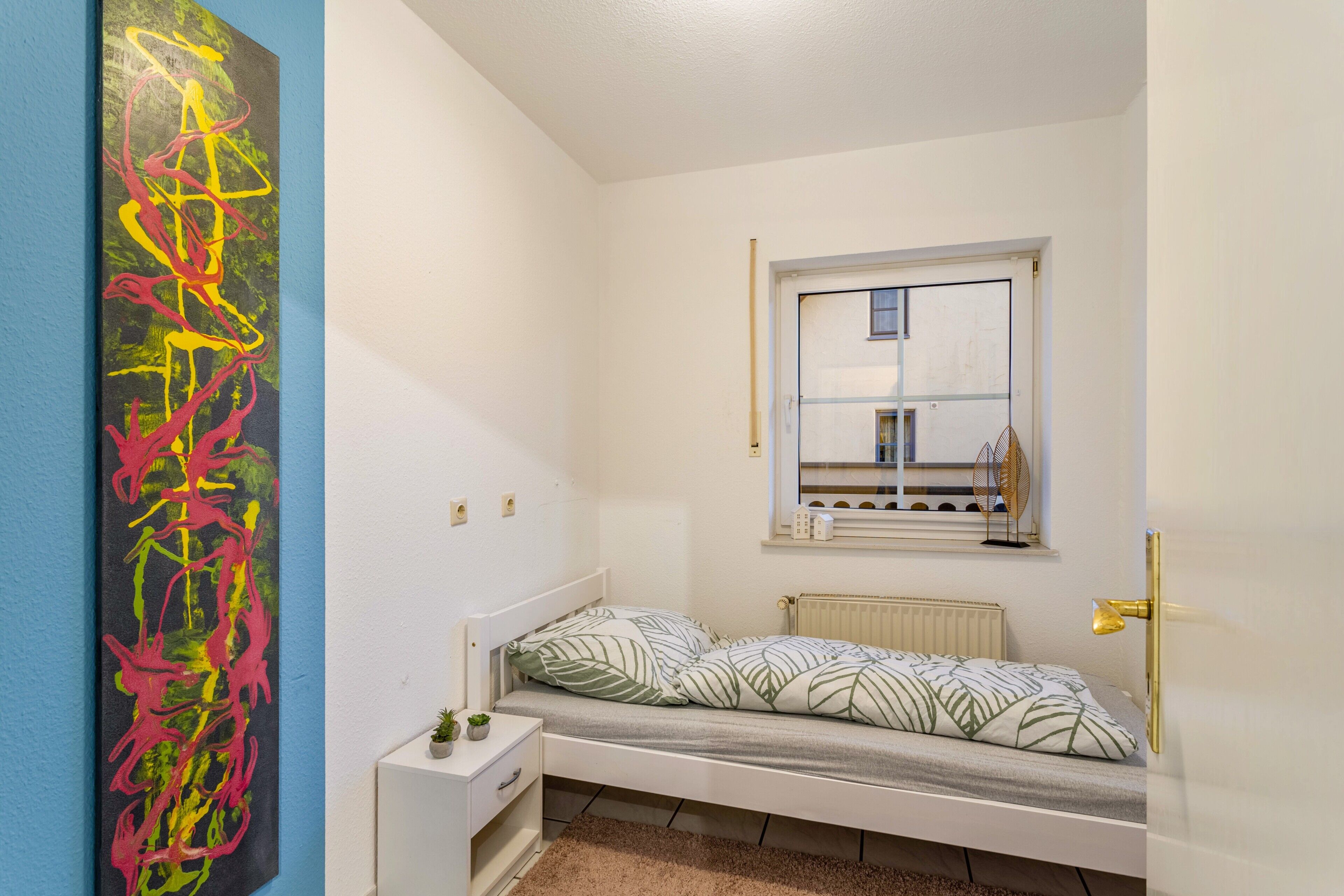 Habitació petita amb un llit individual, una finestra, un radiador, una taula de nit amb una planta i una paret amb obra d'art abstracta.