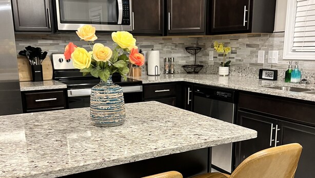 A kitchen with dark cabinets, a granite countertop, a stainless steel microwave, and a vase with yellow flowers.
