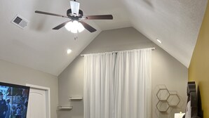A bedroom with a bed, a ceiling fan, a TV, a window with curtains, and a wall-mounted light fixture.