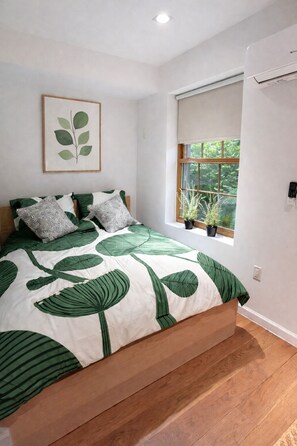 Ein Schlafzimmer mit einem Bett, einem Fenster mit Pflanzen und einer Wand-Klimaanlage.